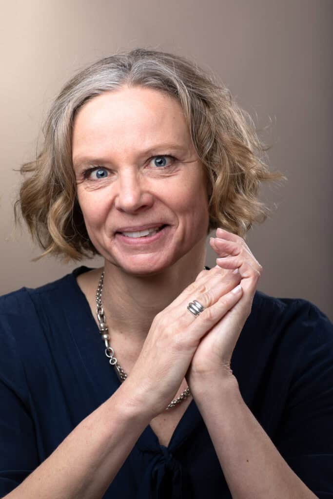 Portrait buste de Colette Constantin, experte en marketing & Communication pour PME. Elle sourit avec les mains paume contre paume devant elle et avec un collier en métal