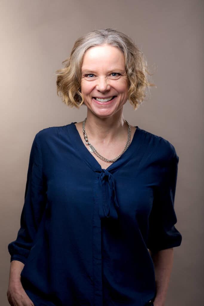 Portrait taille de Colette Constantin, experte en marketing & Communication pour PME. Elle sourie avec les mains le long du corps et avec un collier en métal